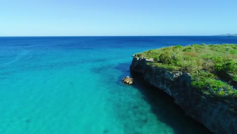 Drohne-Steigt-Entlang-Der-Felsigen-Küste-Hinab-Zum-Klaren,-Blau-türkisen-Wasser-Von-Curaçao