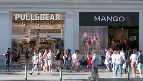Large-crowds-of-pedestrians-and-shoppers-are-seen-walking-past-the-Spanish-multinational-clothing-design-retail-company-by-Inditex,-Pull-and-Bear,-and-Mango-stores-in-Spain