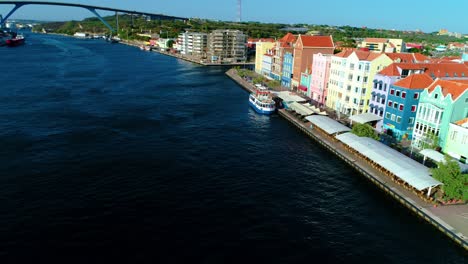 Drone-pullback-aerial-reveals-ferry-and-iconic-Willemstad-world-heritage-buildings-lining-bay,-Queen-Juliana-bridge