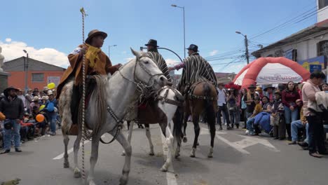 Experience-a-cultural-parade-moment-as-two-riders-elegantly-mounted-on-horses,-wearing-ponchos-and-hats,-proceed-along-the-street-following-the-guidance-of-an-ACOCHA-authority-figure
