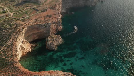 Aerial-orbit-view-of-a-boat-departing-the-rugged-cave-formations-on-the-coastline-near-the-famous-Blue-Lagoon-with-its-crystal-clear-turquoise-waters,-Comino-Island-in-Malta