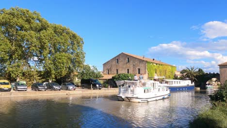 Canal-Du-Midi-at-Le-Somail-France-tourist-boat-passing-through-the-village-and-under-the-bridge-on-a-warm-summer-evening
