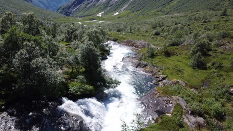 Drone-Siguiendo-Un-Río-Pasando-Por-Algunos-árboles-Con-Los-Fiordos-Nórdicos-Al-Fondo