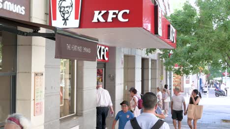 Pedestrians-walk-past-the-American-fast-food-chicken-restaurant-chain,-Kentucky-Fried-Chicken-and-its-logo-in-Spain