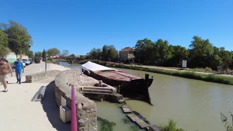 Le-Canal-Du-Midi-Frankreich-Touristen-Genießen-Einen-Spaziergang-Entlang-Des-Kanals-In-Ventenac-En-Minervois-Mit-Einem-Lastkahn,-Der-An-Einem-Sehr-Heißen-Sommertag-Am-Kanal-Festgemacht-Hat