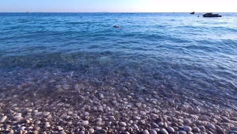 Seawater-washing-white-pebbles-on-beautiful-beach,-boy-snorkeling-and-motorboats-floating-on-calm-water-in-Mediterranean