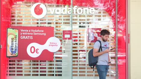 A-man-using-a-smartphone-stands-outside-a-closed-British-multinational-telecommunications-corporation-and-phone-operator,-Vodafone,-store-in-Spain