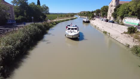 Canal-Du-Midi-at-Ventenac-En-Minervois-as-a-canal-boat-passes-under-a-bridge-in-the-village-on-a-warm-summer-day