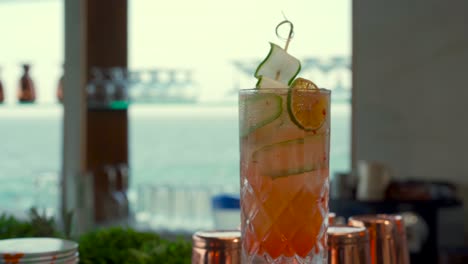 Tequila-sunrise-mezcal-cucumber-an-lemon-garnish-cocktail-bayside-bayfront-fresh-drink-beach-refresher-sea-background-blurry