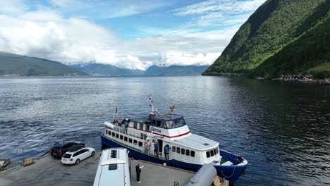 Tourist-sightseeing-boat-departing-from-Vik-in-Sogn-Norway---Summer-cruise-with-fjord-landscape-background-and-bus-left-on-quay-with-busdriver-waving-his-hand