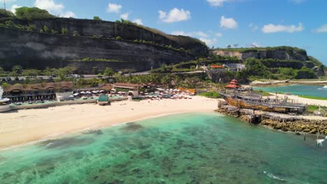 Minoo-Beach-Club-White-Sand-Shore-with-Turquoise-Calm-Sea-in-Uluwatu-Bali,-Melasti-Beach-with-Steep-Cliffs-in-Background---Aerial-pushback-shot