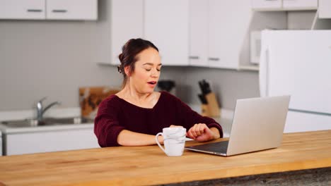 A-Young-Woman-Sitting-at-her-Home-Kitchen-Counter-with-a-Cup-of-Coffee-Opens-a-Laptop-and-Smiles-Typing-and-Browsing-the-Internet