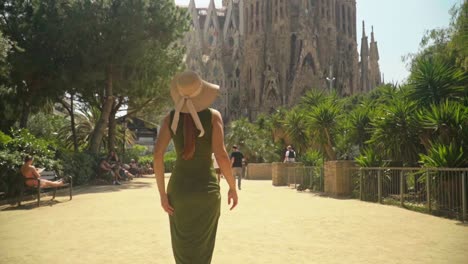 Stunning-video-of-a-young-caucasian-female-in-a-green-dress-with-a-knitted-hat,-walking-to-incredible-landmark-building---Sagrada-Familia,-Barcelona