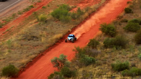 Off-Road-Buggy-Racing-On-Red-Dirt-Outback-Track-Alongside-Rural-Highway-In-Alice-Springs,-Australia,-4K-Telephoto-Drone