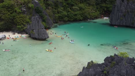 Tourists-kayaking-and-swimming-in-tropical-emerald-Cadlao-Lagoon,-El-Nido,-Karst-rock-formations-at-Island,-Philippines,-Aerial-view