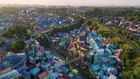 Bird-eye-view-of-Jodipan-Village-aka-Rainbow-Village-with-a-train-line-passing-next-to-it-and-a-bridge-with-cars-passing-on-it---sunset-time-in-Malang,-East-Java---Indonesia