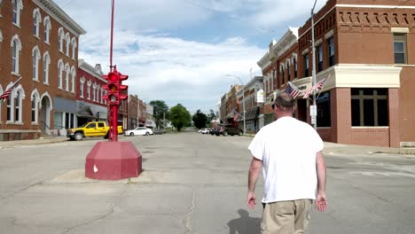 Man-in-white-shirt-and-sunglasses-walking-towards-an-antique-four-way-stop-light-in-downtown-Toledo,-Iowa-with-gimbal-video-following-from-behind