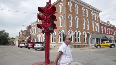 Man-in-white-shirt-and-sunglasses-sitting-at-the-base-of-an-antique-four-way-stop-light-in-downtown-Toledo,-Iowa-with-video-moving-around-from-behind