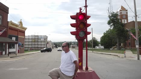 Man-in-white-shirt-and-sunglasses-sitting-at-the-base-of-an-antique-four-way-stop-light-in-downtown-Toledo,-Iowa-with-video-moving-around