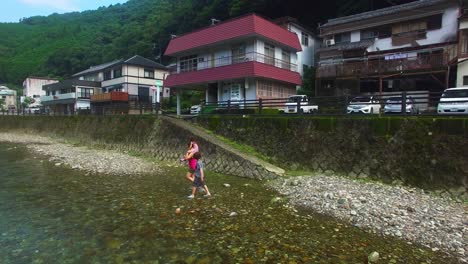 Drone's-eye-view-captures-a-serene-moment-in-Koyasan-village,-Japan,-as-a-mother-with-2-children-traverse-the-river-on-foot,-connected-by-its-tranquil-embrace
