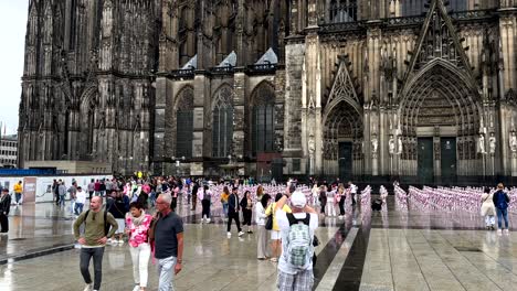 Muchos-Turistas-Visitan-La-Catedral-De-Colonia-Con-Obras-De-Arte-Al-Aire-Libre-Durante-El-Día-Lluvioso,-Alemania---Cámara-Lenta---Tema-De-Explotación-Infantil-Con-Muñecas-Rosas