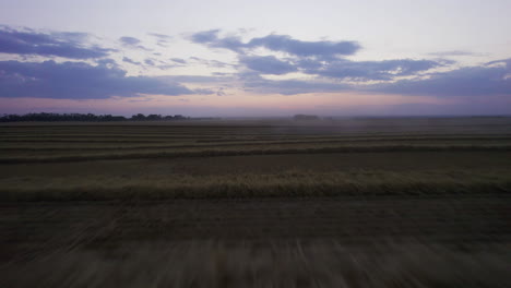 Fast-low-aerial-pullback-over-harvested-wheat-fields-with-combine,-twilight