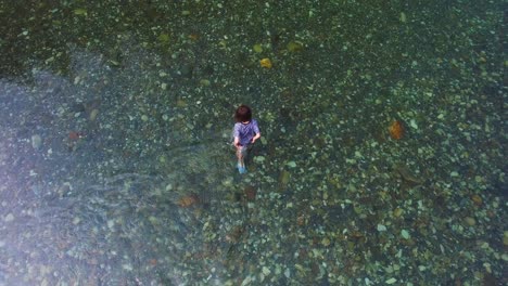 Drone's-high-angle-captures-a-young-male-explorer-in-a-clear-river,-revealing-the-stunning-riverbed