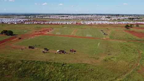 Drone-approach-over-kids-walking-onto-football-field,-wide-landscape-with-buildings-in-the-background---Posadas-hipodrome