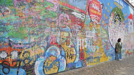 Woman-stands-in-front-of-John-Lennon-Wall-In-Prague,-Czech-Republic---Inspired-Graffiti,-Lyrics-From-Beatles'-Songs,-And-Designs-Relating-To-Local-And-Global-Causes