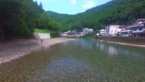 Aerial-drone-glides-over-serene-Japanese-village-Koyasan,-capturing-man-walking-along-the-shore-of-a-tranquil-river