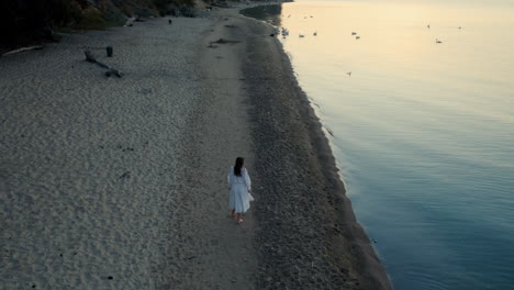 Cinematic-drone-shot-of-beautiful-woman-in-white-dress-walking-alone-on-lonely-beach
