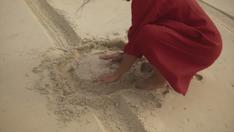 Anzeige-Erwachsenes-Mädchen-In-Einem-Roten-Kleid,-Das-Barfuß-Mit-Sand-Am-Strand-Spielt