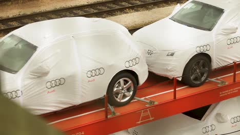 Handheld-camera-shot-of-an-orange-train-car-with-the-Audi-logo-carrying-two-Audi-cars,-veiled-in-white-protective-cover,-amidst-a-backdrop-of-train-tracks-and-other-cars
