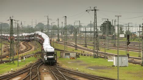 Handheld-camera-catches-a-white-freight-train-with-a-blue-stripe-curving-around-a-bend-amidst-a-busy-train-yard,-multiple-tracks-and-trains,-electrical-poles-under-an-overcast-sky