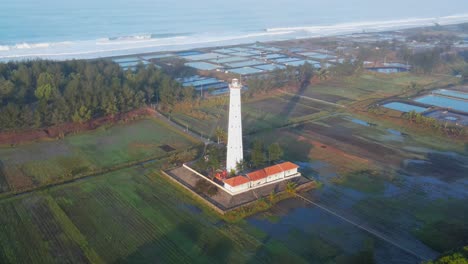 Aerial-orbit-shot-of-lighthouse-near-beach-and-Ocean-in-Central-Java,-Indonesia---Agricultural-plantaiton-fields-in-the-morning