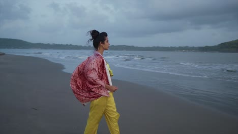 Cinematic-shot-of-a-Beautiful-indian-asian-model-woman-walking-along-tropical-beach-with-colorful-swimsuit-on-a-cloudy-evening