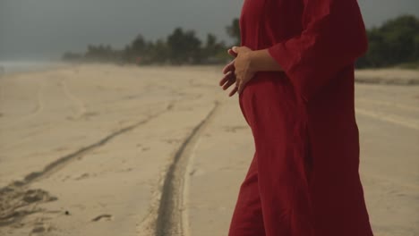 girl-in-a-red-dress-stands-on-a-beach-and-puts-her-hands-in-her-red-dress's-pocket-on-a-sunny-day