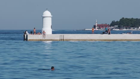 People-sun-bathing-at-Zadar-promenade-with-beacon-and-distant-lighthouse,-Croatia-summer