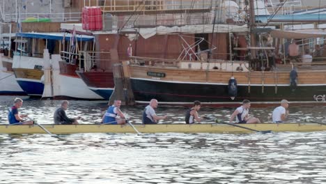 Nine-rowers-passing-docked-ships-in-port-of-Zadar-old-town,-Croatia