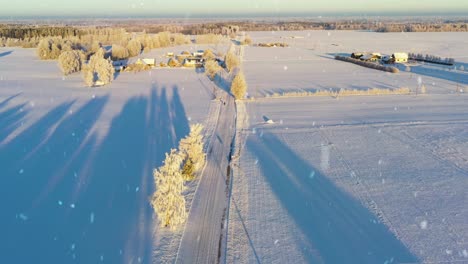 Snow-covered-agriculture-fields-and-small-village-with-homes-during-snowfall,-aerial-view