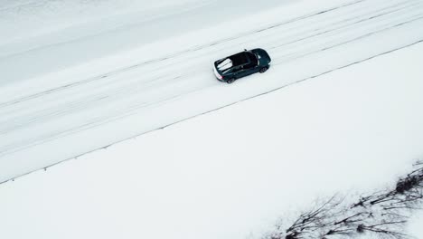 an-electric-car-is-driving-on-a-snow-covered-road-in-the-arctic-circle-during-a-snowfall,-followed-by-a-drone