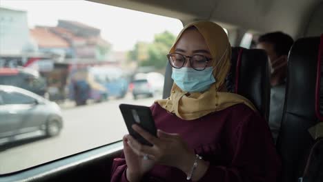 Positive-cheerful-young-woman-in-casual-clothes-sitting-in-car-center-seat-wearing-mask,-glasses,-headscarf-and-looking-away-after-using-mobile-phone