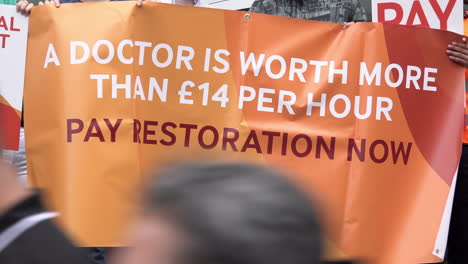 An-orange-banner-that-reads,-“A-doctor-is-worth-more-than-£14-per-hour,-pay-restoration-now”-is-held-by-striking-Junior-Doctors-on-a-picket-line-outside-the-University-College-Hospital