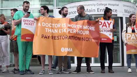 Striking-Junior-Doctors-stand-on-a-picket-line-outside-the-University-College-Hospital-holding-an-orange-banner-that-reads,-“A-doctor-is-worth-more-than-£14-per-hour,-pay-restoration-now”