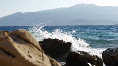 Close-up-of-large-rocks-on-the-Albanian-beach-being-shattered-by-the-blue-waves-of-the-Ionian-Sea,-with-beautiful-mountains-in-the-background