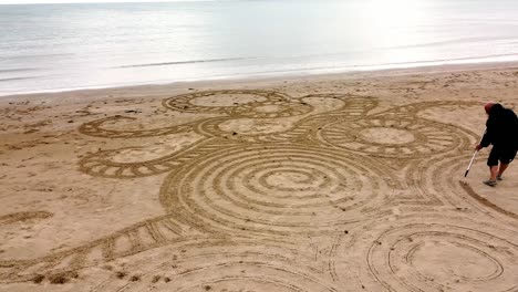Aerial-view-circling-man-creating-zen-sand-art-design-on-windy-Welsh-golden-beach