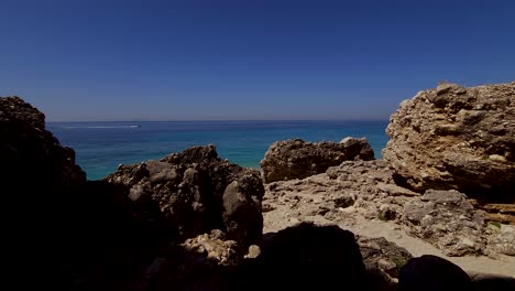 Rocky-seaside-in-Mediterranean-blue-turquoise-sea,-jet-ski-in-horizon-seen-through-cliffs-at-the-quiet-beach