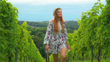 Stunning-HD-footage-of-a-young-white-Caucasian-woman-with-a-knitted-hat,-dress-and-red-lipstick-walking-through-vineyards-with-a-bottle-of-wine-and-a-glass-in-hand