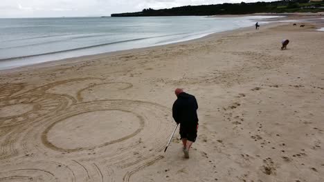 Aerial-view-circling-man-creating-sand-art-design-on-windy-Welsh-golden-beach