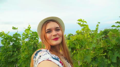 Stunning-HD-footage-of-a-young-white-Caucasian-woman-with-a-knitted-hat-and-red-lipstick-holding-the-camera,-smiling-and-posing-among-the-vineyards